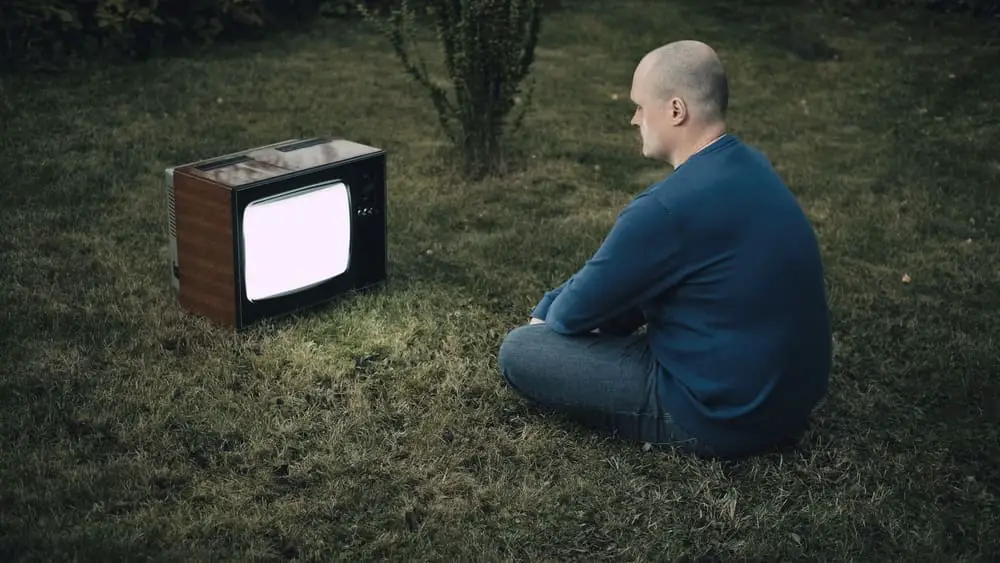 Bald man is sitting on grass and looking at old retro TV. TV is strobing